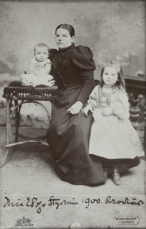 Fotografia siedzącej kobiety. Po lewej stronie, na stoliku siedzi niemowlę, po prawej kilkuletnia dziewczynka przytulona do kolana kobiety